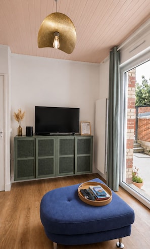 Una sala moderna con televisor de pantalla plana, un mueble verde, un reposapiés azul y un ventanal con vista a un muro de ladrillo y follaje.