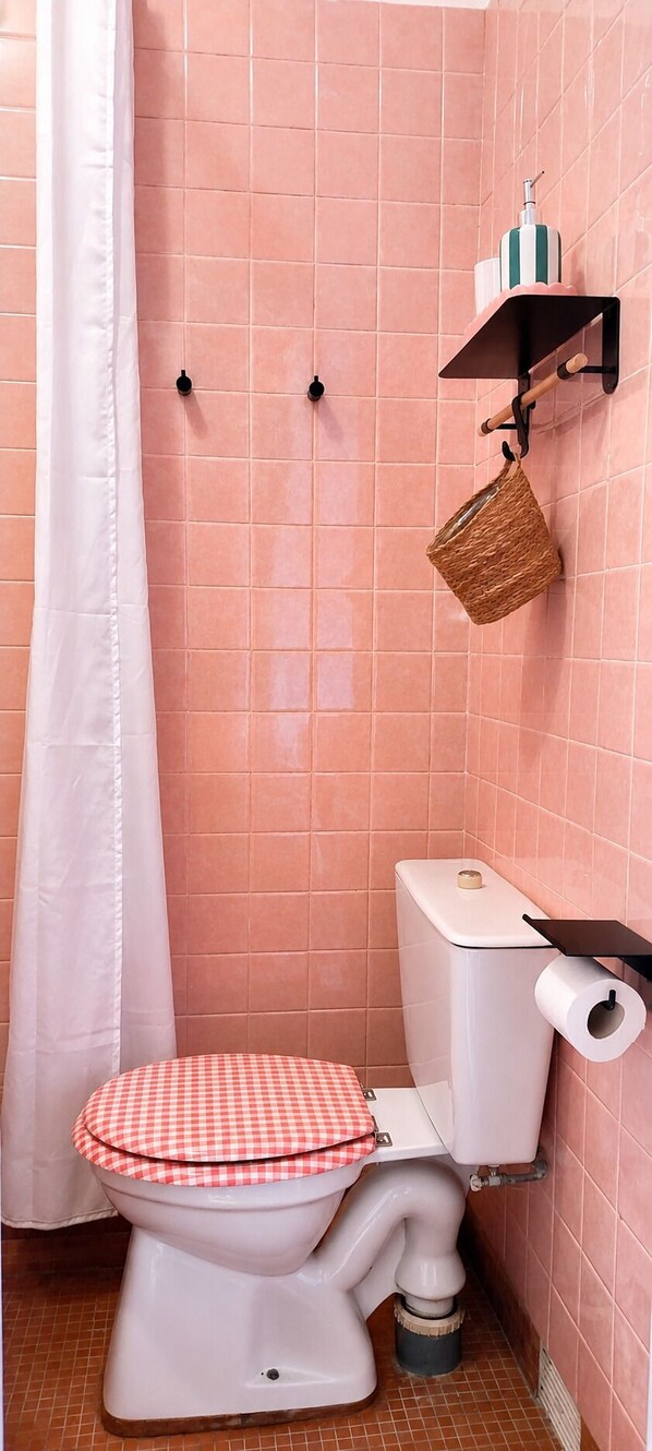 A bathroom with pink tiles, a white toilet with a checkered seat cover, a shower curtain, a hanging basket, and a toilet paper holder.