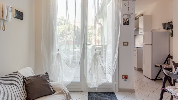 A room with a bed, a chair, a kitchen area, and a door with sheer curtains. - Apartment 'Un Angolo Nel Verde A Empoli' with Shared Terrace, Wi-Fi and Air Conditioning (Empoli)