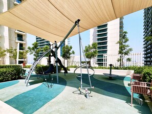 Un terrain de jeux avec des balançoires et un toboggan, entouré de verdure et de bâtiments modernes.