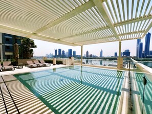 An infinity pool with a city skyline view, lounge chairs, and a covered area with a pergola.
