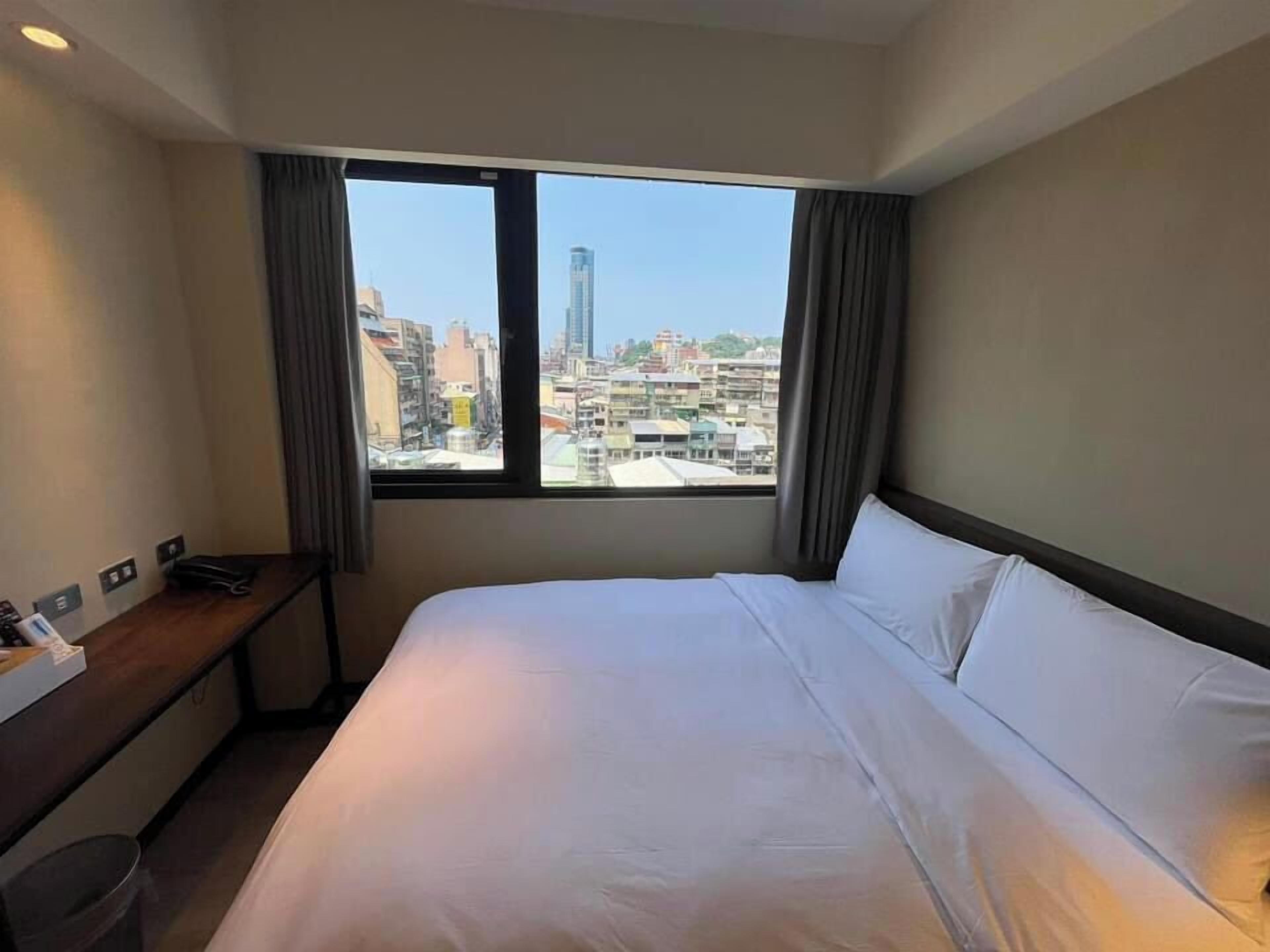 A hotel room with a large bed, a desk, and a view of a cityscape through a window.