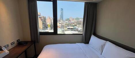 A hotel room with a large bed, a desk, and a view of a cityscape through a window.