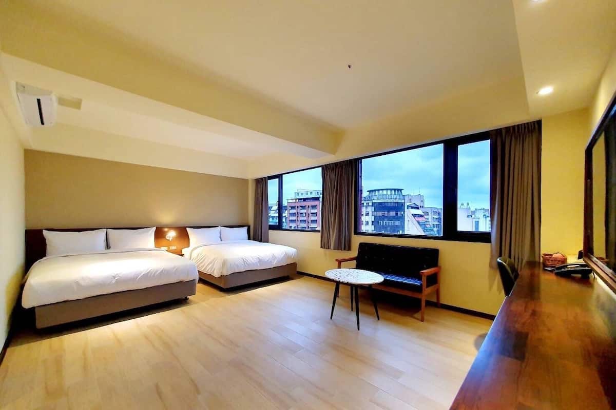 Habitación de hotel con dos camas, una mesita, un sofá y un ventanal con vistas a la ciudad.