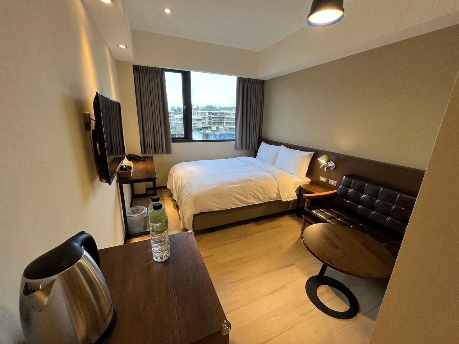 Ein Hotelzimmer mit einem Bett, einem Schreibtisch mit Wasserkocher, einem Sessel, einem an der Wand montierten Fernseher und einem Fenster mit Vorhängen.