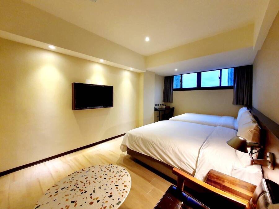 Habitación de hotel con una cama grande, un televisor de pantalla plana en la pared, una mesita redonda y una ventana con cortinas.