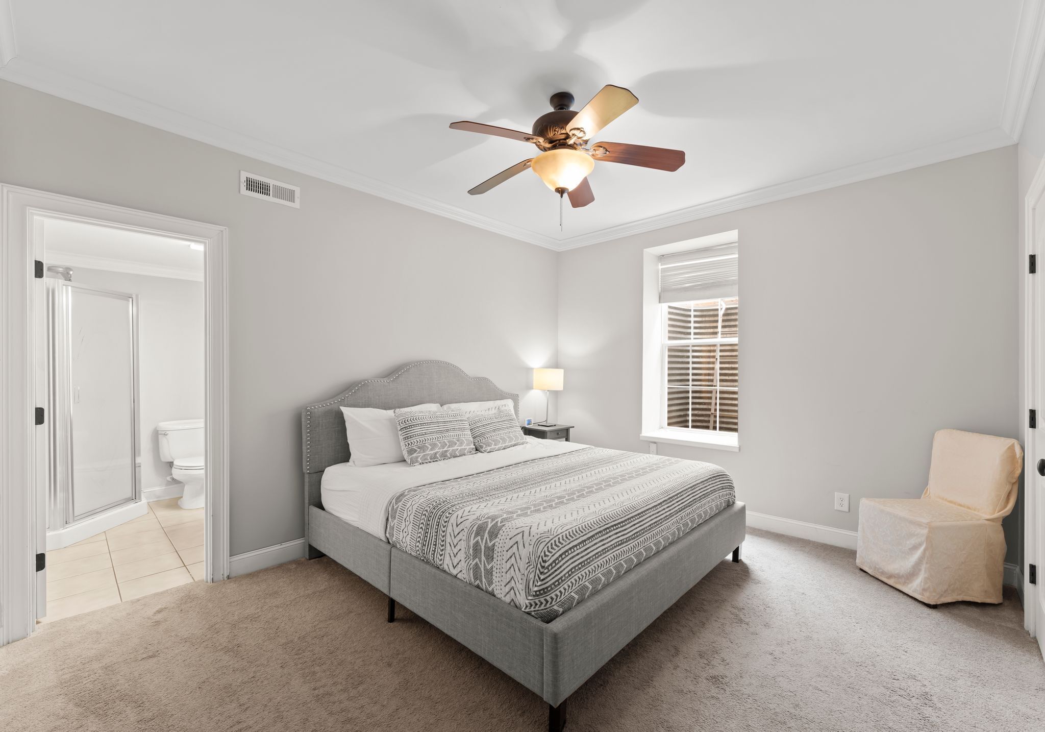 A bedroom with a bed, a ceiling fan, a window with blinds, a chair, and a door leading to a bathroom.