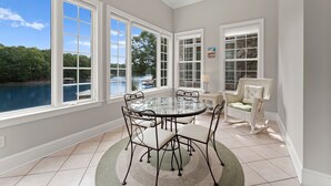 A dining area with a glass table surrounded by chairs, large windows offering a view of a body of water, and a wicker chair by the window. - Sleeps 20! Lakefront + Private Dock + Pool | Whitetail Cove By AvantStay (Troutman)