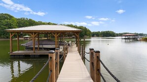 A wooden pier leading to a covered seating area with a boat docked on the left. - Sleeps 20! Lakefront + Private Dock + Pool | Whitetail Cove By AvantStay (Troutman)