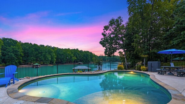 A swimming pool with a stone border, surrounded by a fence, and a bench with an umbrella nearby. - Sleeps 20! Lakefront + Private Dock + Pool | Whitetail Cove By AvantStay (Troutman)