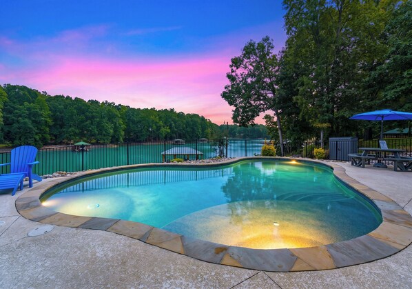 A swimming pool with a stone border, surrounded by a fence, and a bench with an umbrella nearby.