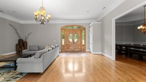 A spacious living room with hardwood flooring, a grey sofa, a coffee table, and a chandelier. - Sleeps 20! Lakefront + Private Dock + Pool | Whitetail Cove By AvantStay (Troutman)