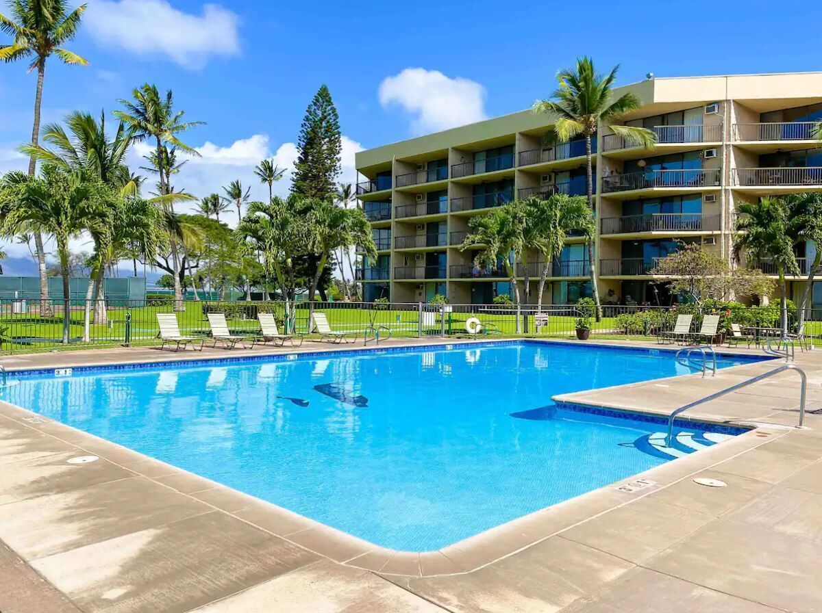 Piscina com espreguiçadeiras, rodeada por palmeiras e um edifício com várias andares e varandas.