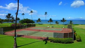 Ein Tennisplatz mit roter Belag, umgeben von grünem Rasen und Palmen, mit blauem Himmel und Blick auf den Ozean im Hintergrund.