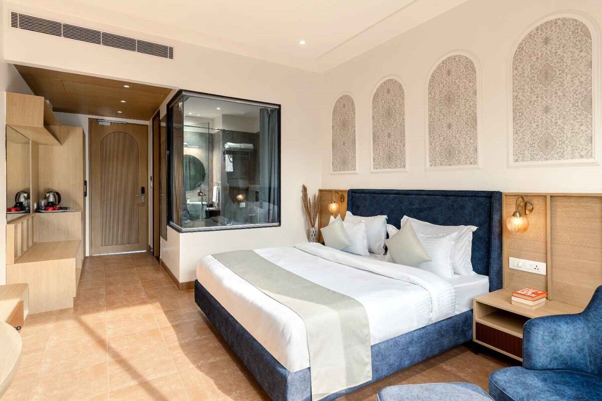 Una habitación de hotel moderna con una cama grande, una zona de estar y vistas al baño a través de una puerta de vidrio.