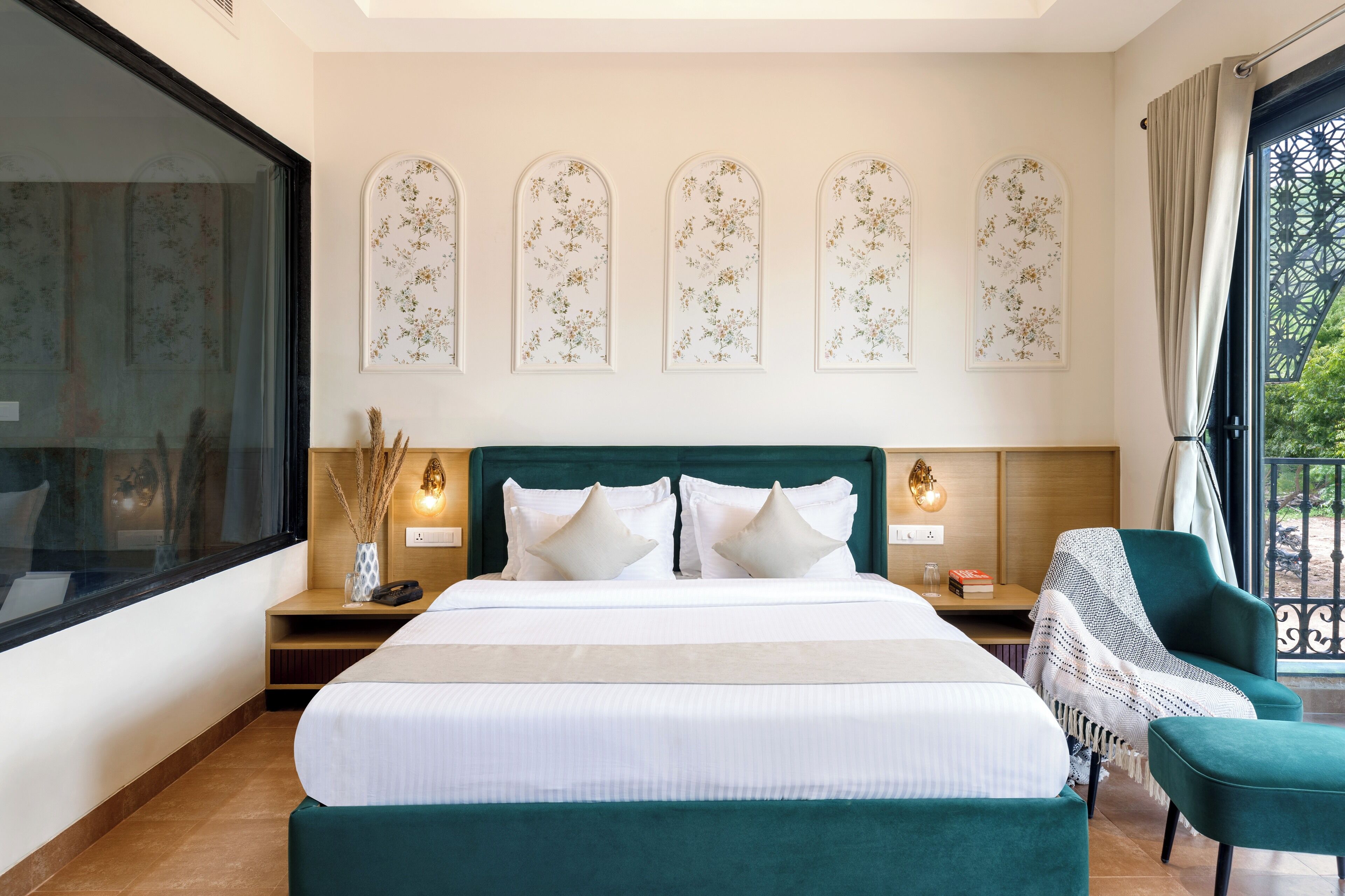 Habitación de hotel con una cama grande, una mesita de noche con lámpara, una silla y vistas a zonas verdes por la ventana.
