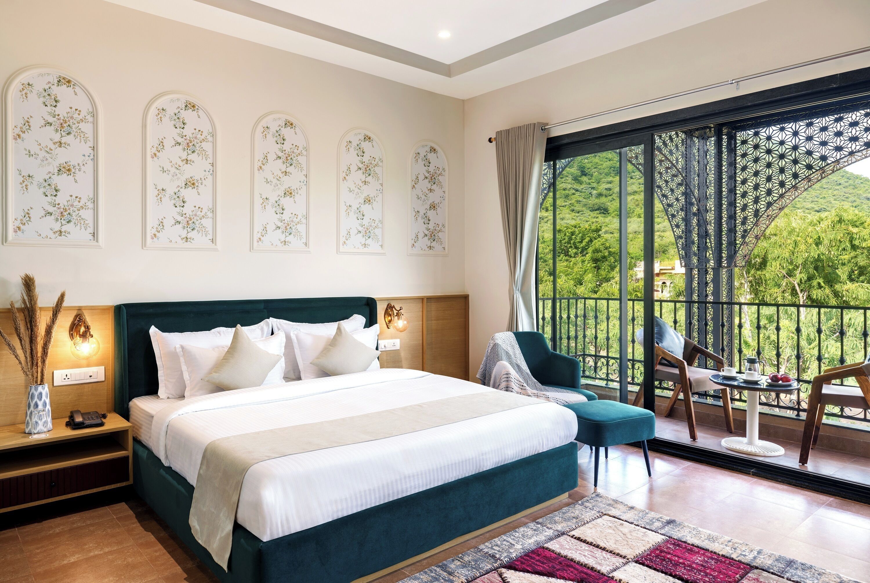 A bedroom with a large bed, a nightstand, a bedside table with a lamp, a chair, a small table with a cup and saucer, and a balcony with a view of greenery.