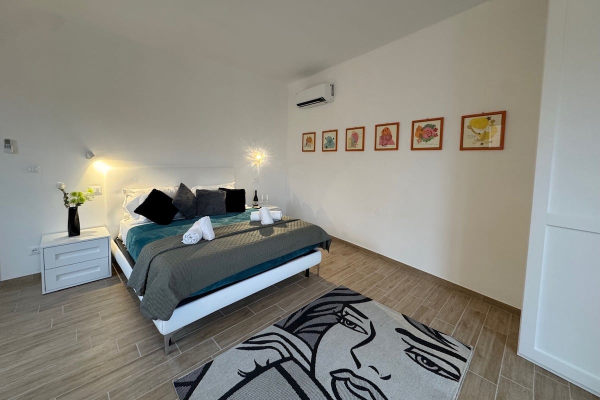 Un dormitorio con cama, mesitas de noche, un florero con flores y decoración en la pared.