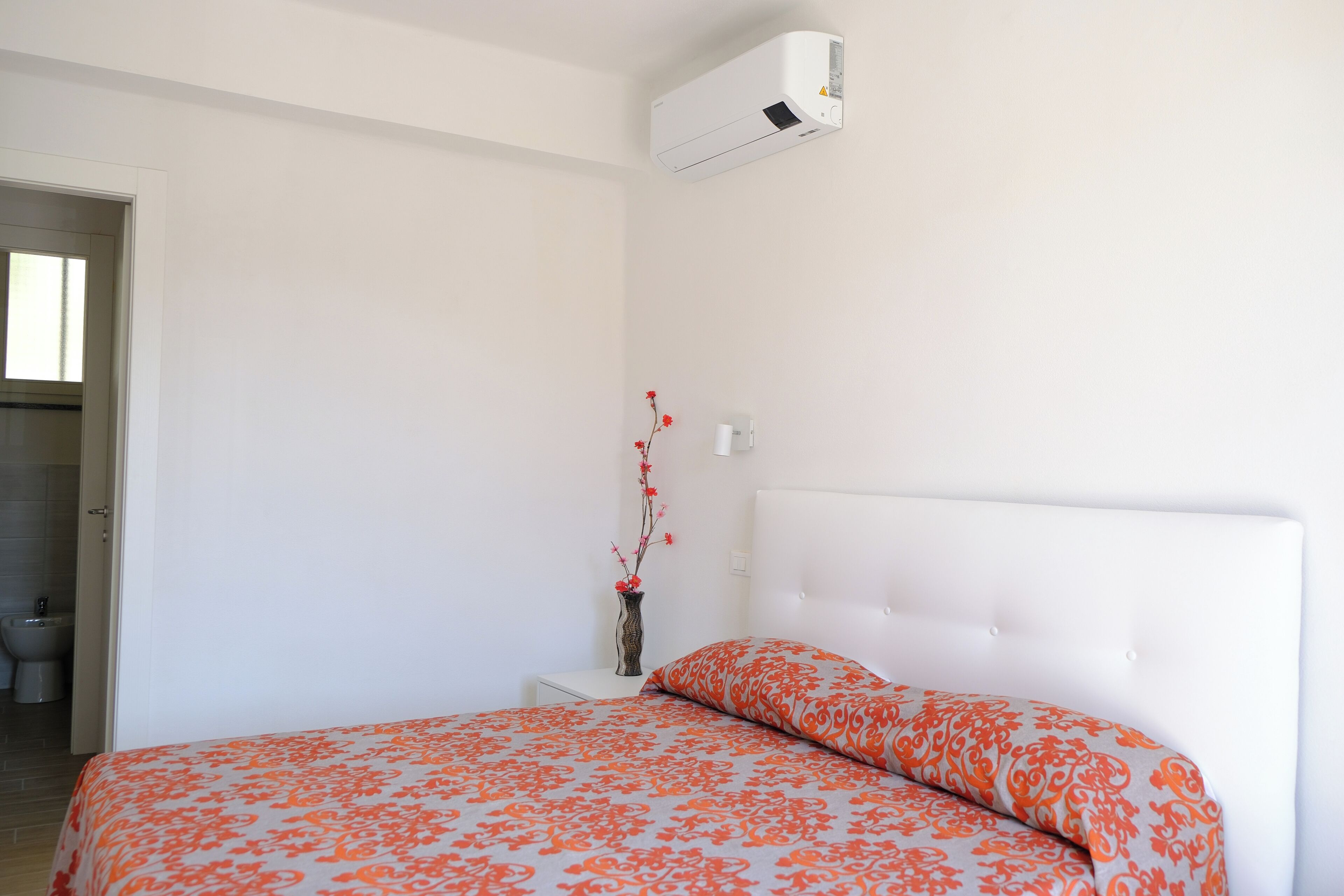 Un dormitorio con una cama, una mesita de noche, un florero con flores y un equipo de aire acondicionado.