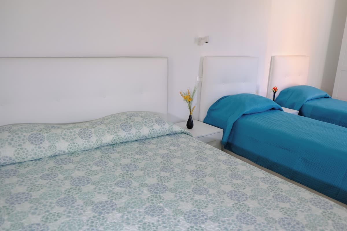 Un dormitorio con dos camas, una con cubrecama estampado y la otra con cubrecama azul liso. Entre las camas hay una mesita de noche con un jarrón de flores.