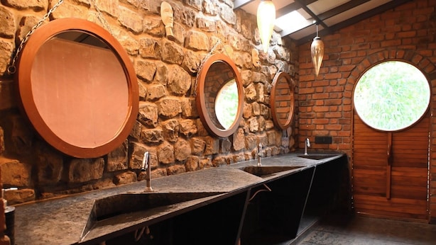 A restroom with three sinks, each with a round mirror and a wooden frame. The walls are made of exposed stone, and there is a window on the right wall. - Bamboo and Bricks Resorts (Paud)