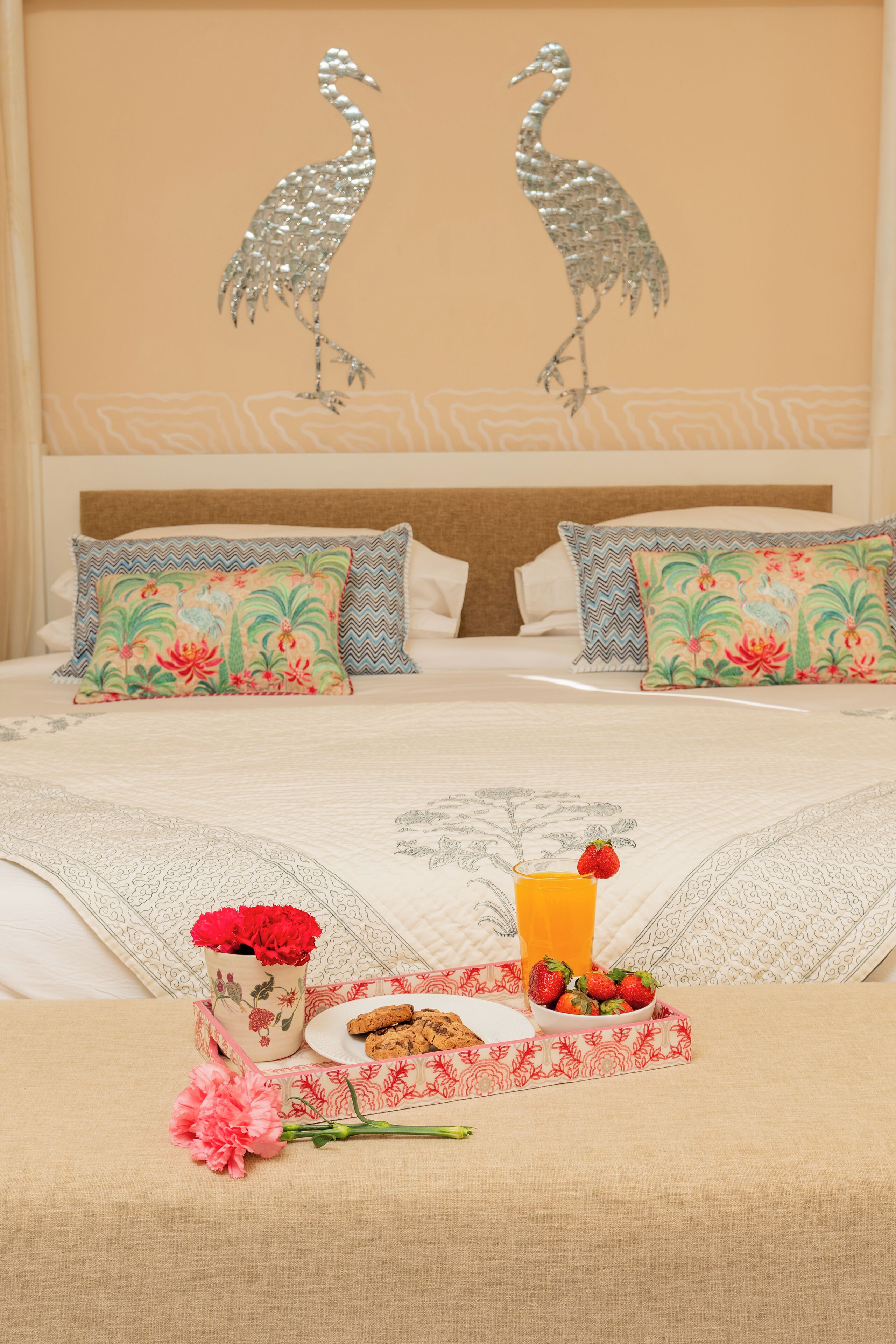 Una cama bien hecha con almohadones florales, un adorno en la pared y una bandeja de desayuno con galletas, fresas y zumo de naranja.