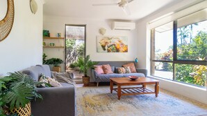 A living room with a grey sofa, a wooden coffee table, a patterned rug, and a large window with a view of greenery. - Tropical Treasure | Outdoor Kitchen & Shower (Wanguri)