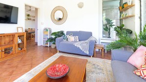 A living room with a sofa, coffee table, and a TV. There are plants and shelves with items on the walls. - Tropical Treasure | Outdoor Kitchen & Shower (Wanguri)