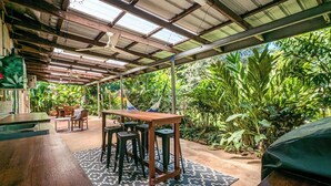 A covered outdoor area with a wooden table, four stools, and a patterned rug. - Tropical Treasure | Outdoor Kitchen & Shower (Wanguri)
