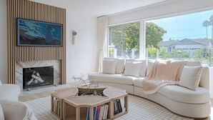 A modern living room with a fireplace, a large window, and a white sofa. - 4-bed designer family-friendly home with large yards & theater room near beaches (Los Angeles)