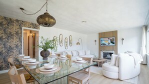 A modern living room with a glass dining table, white sofas, and a fireplace. - 4-bed designer family-friendly home with large yards & theater room near beaches (Los Angeles)