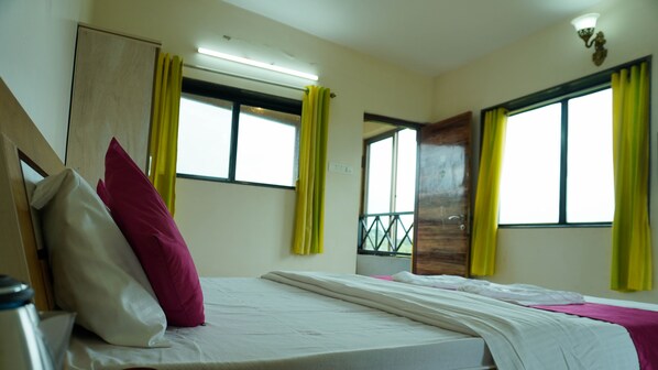 A hotel room with two beds, a wooden wardrobe, a window with yellow curtains, and a door with a metal grille. - Forest Leaf (Mahabaleshwar)