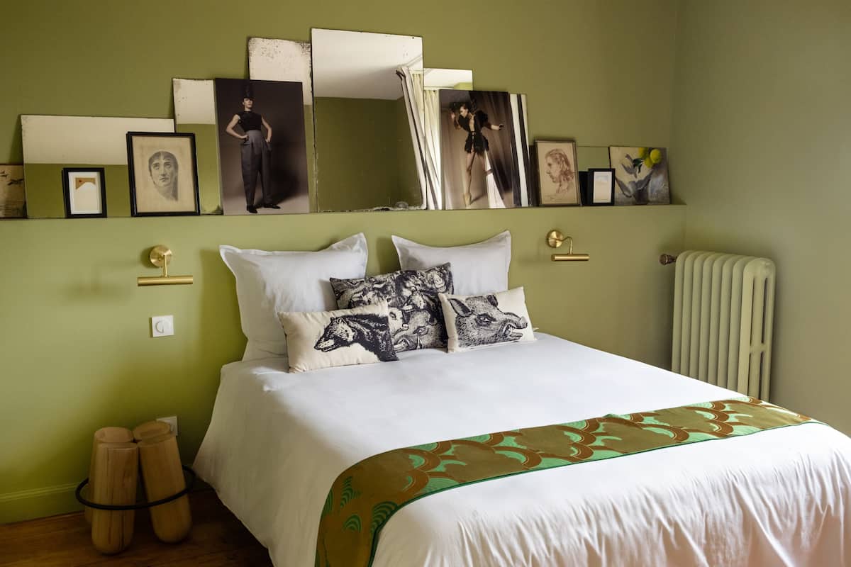 A neatly made bed with white linens and patterned pillows, a bedside table with a lamp, and a wall-mounted mirror above the bed.