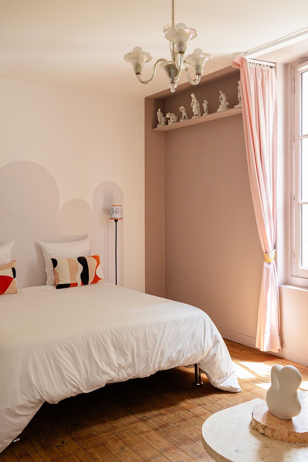 A bedroom with a bed, a round table, a sculpture, a shelf with figurines, and a chandelier.