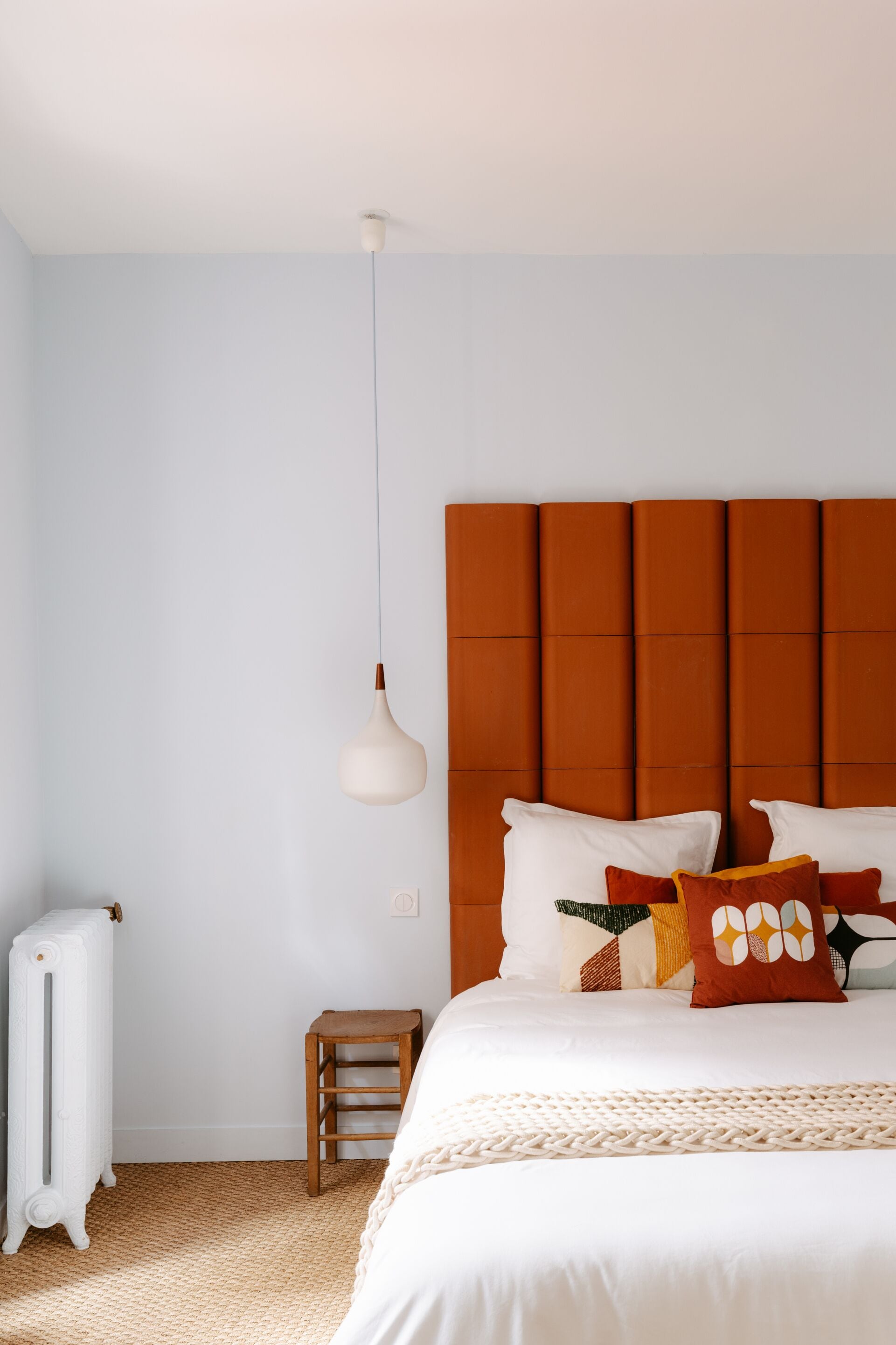 Een slaapkamer met een wit bed, een houten nachtkastje, een radiator en een hanglamp.