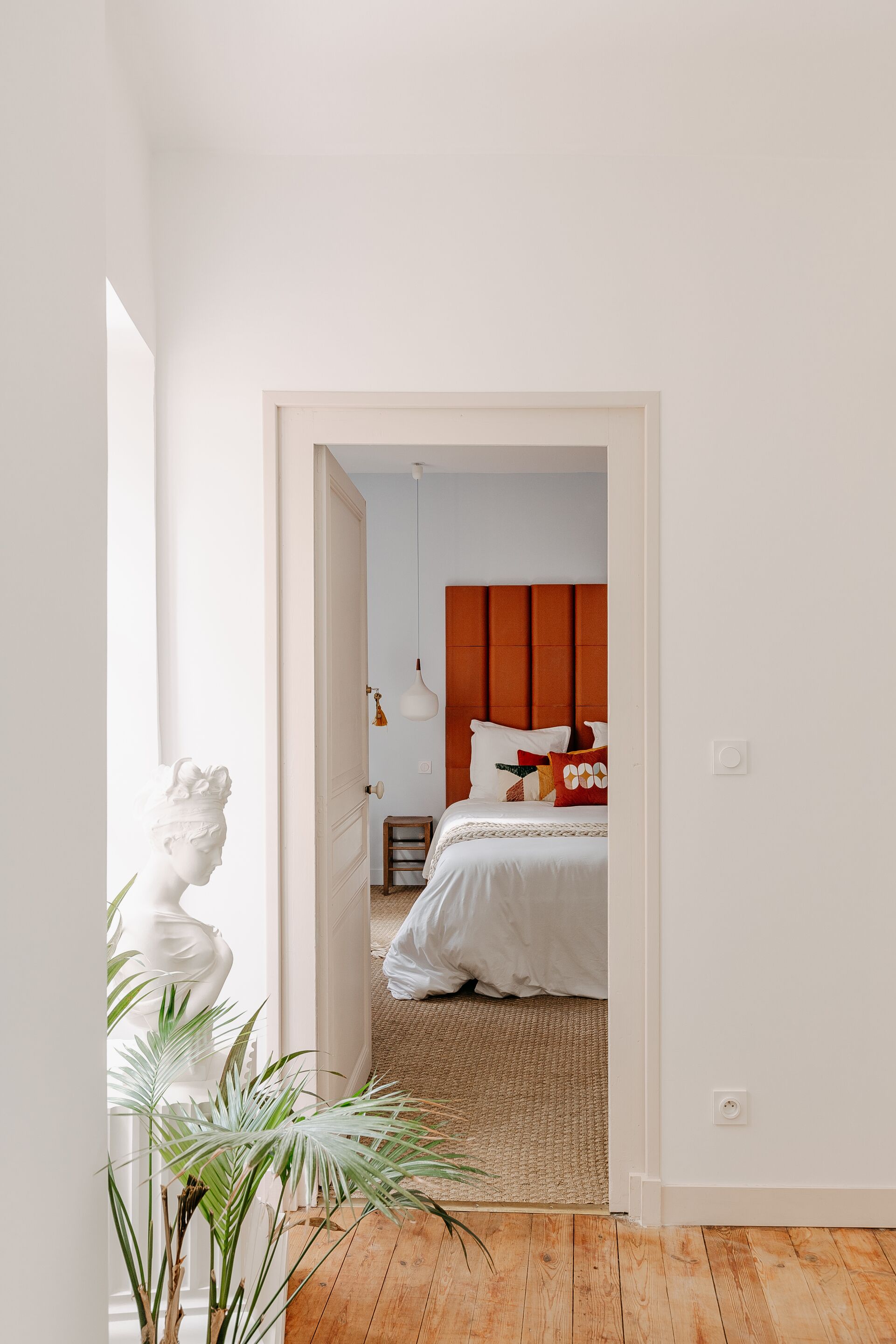 Une chambre avec un plancher en bois, un lit avec une literie blanche, une tête de lit rouge et une sculpture en buste blanche.