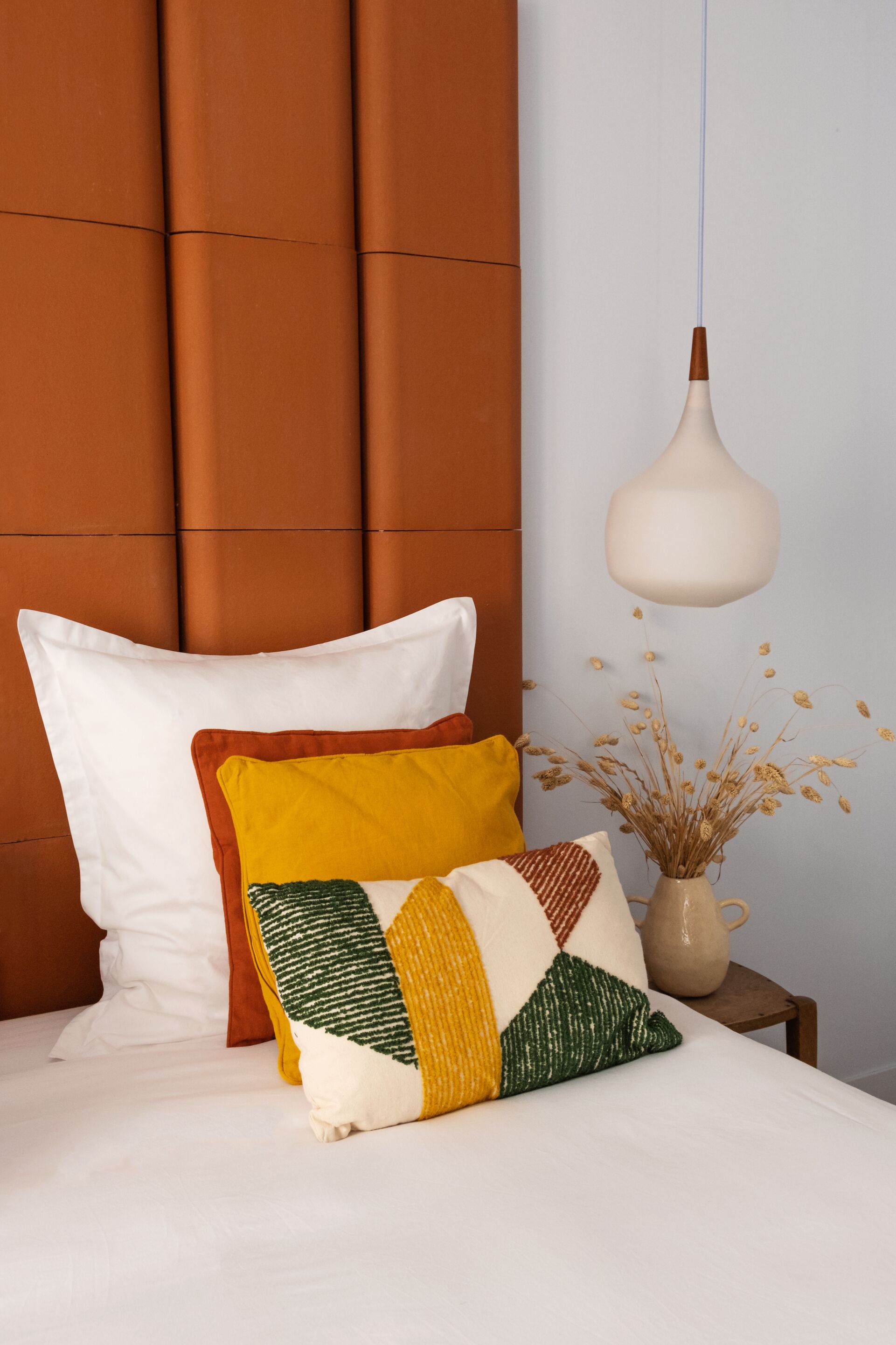 Una cama con almohadas blancas y de varios colores, un florero con plantas secas y una lámpara colgante.