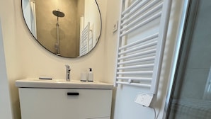 A bathroom with a white vanity, a round mirror, a towel rack, and a glass shower door. - Cosy apartment 1 min from Paris (Charenton-le-Pont)