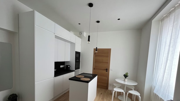 A modern kitchen with white cabinets, a black countertop, and a wooden door. There is a small round table with chairs and a potted plant. - Cosy apartment 1 min from Paris (Charenton-le-Pont)