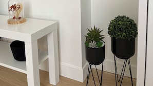 A white wall with a mounted TV, a small white table with a decorative item, and two black potted plants on metal stands. - Cosy apartment 1 min from Paris (Charenton-le-Pont)