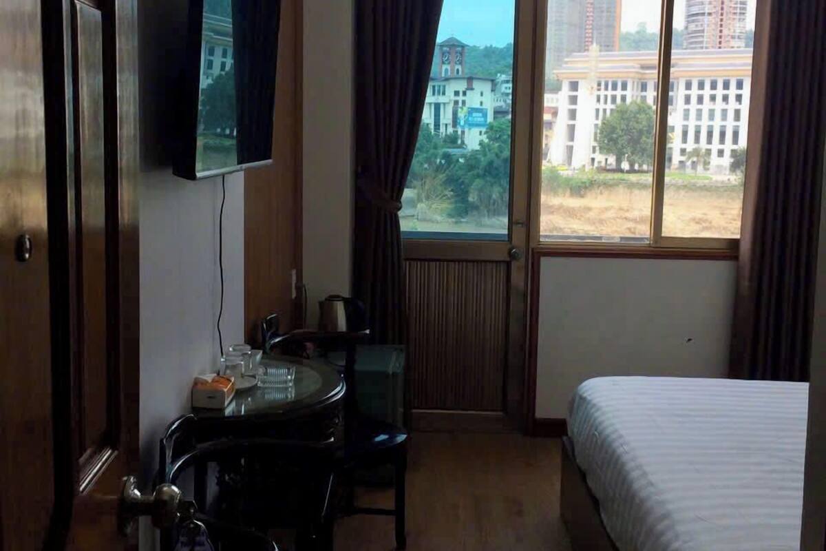 A hotel room with a bed, a desk with a chair, a television mounted on the wall, and a window with curtains.