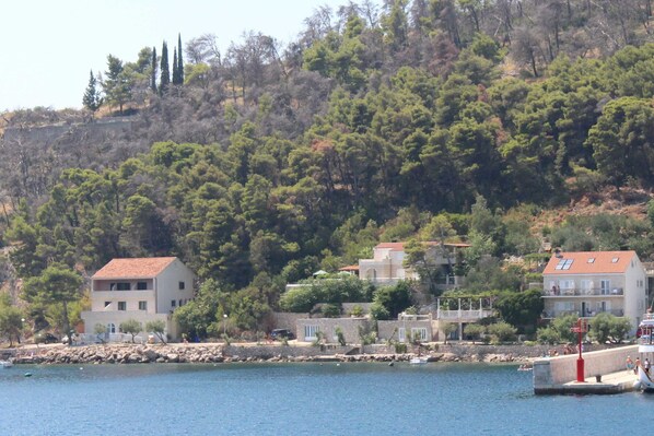 A coastal building complex with multiple structures, including a two-story house and a larger building with a red roof, situated on a hillside with dense trees. - 2 bedroom apartment with Sea view (Trstenik)