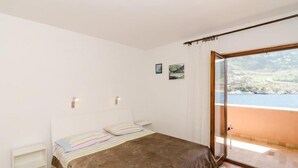 A bedroom with a bed, a small bedside table, a nightstand, a window with a view of the sea, and a door leading to an outdoor balcony. - Apartments Bezek - Studio Apartment with Sea View (Trstenik)