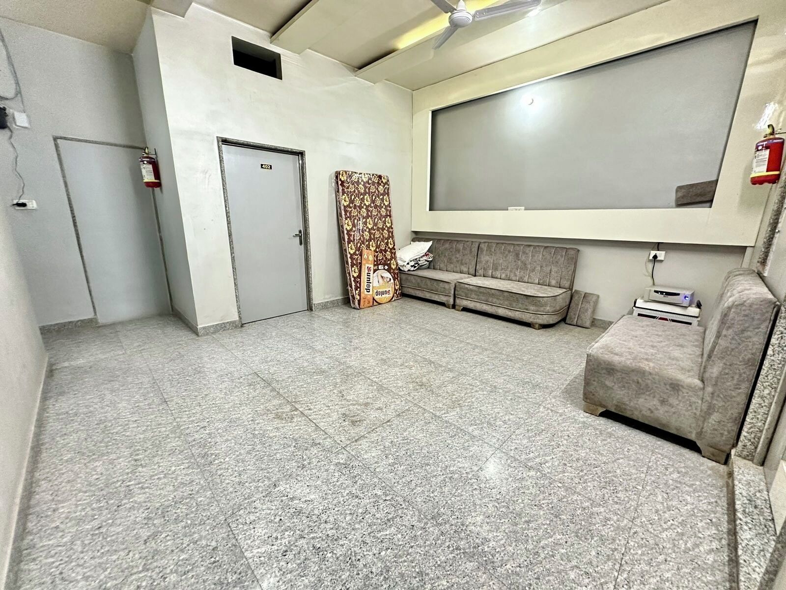 A room with a grey sofa, a small table with a device, a patterned curtain, and a door marked with the number 402.