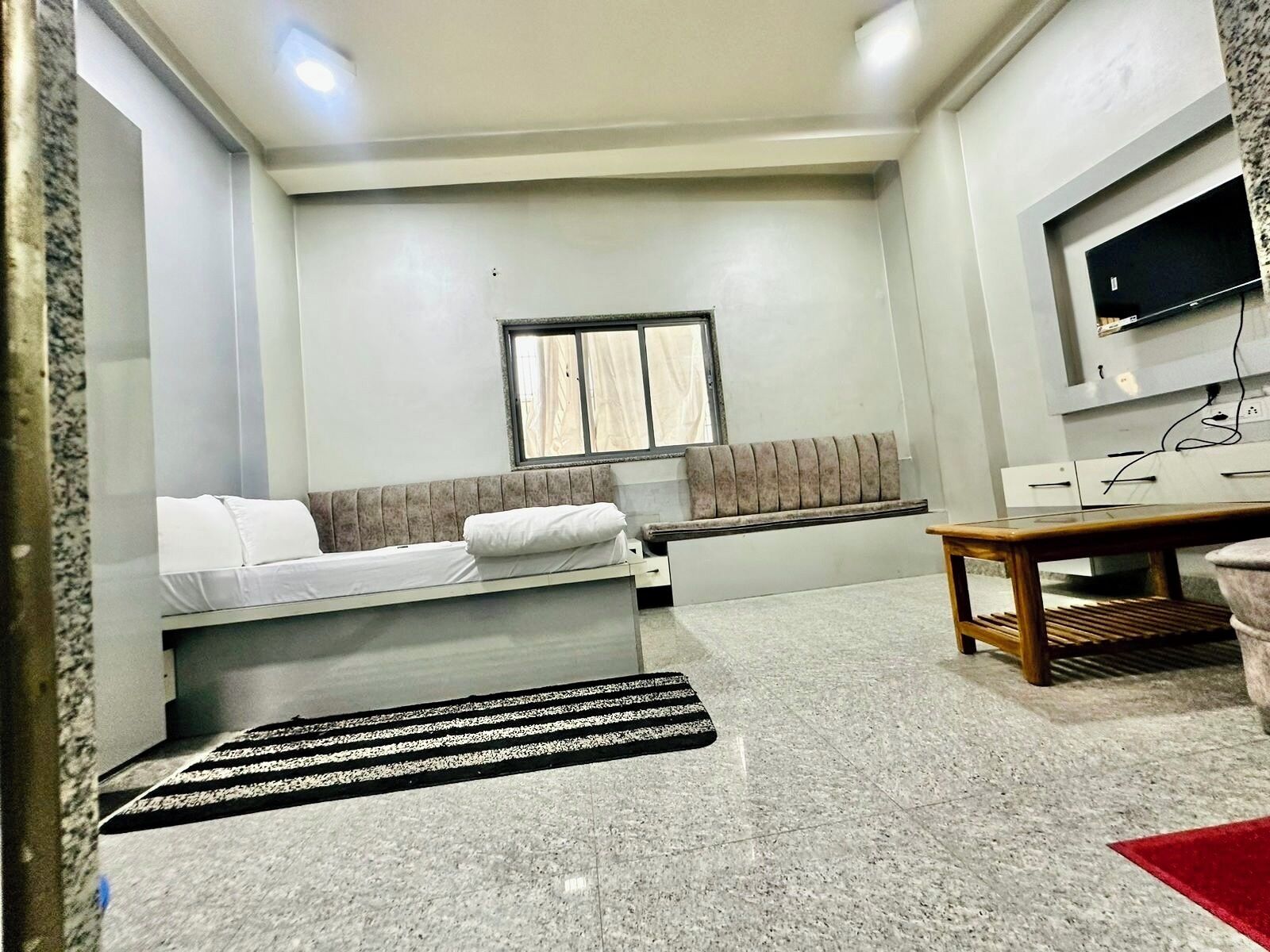 A compact room with a bed, a bench, a small table, and a TV mounted on the wall.