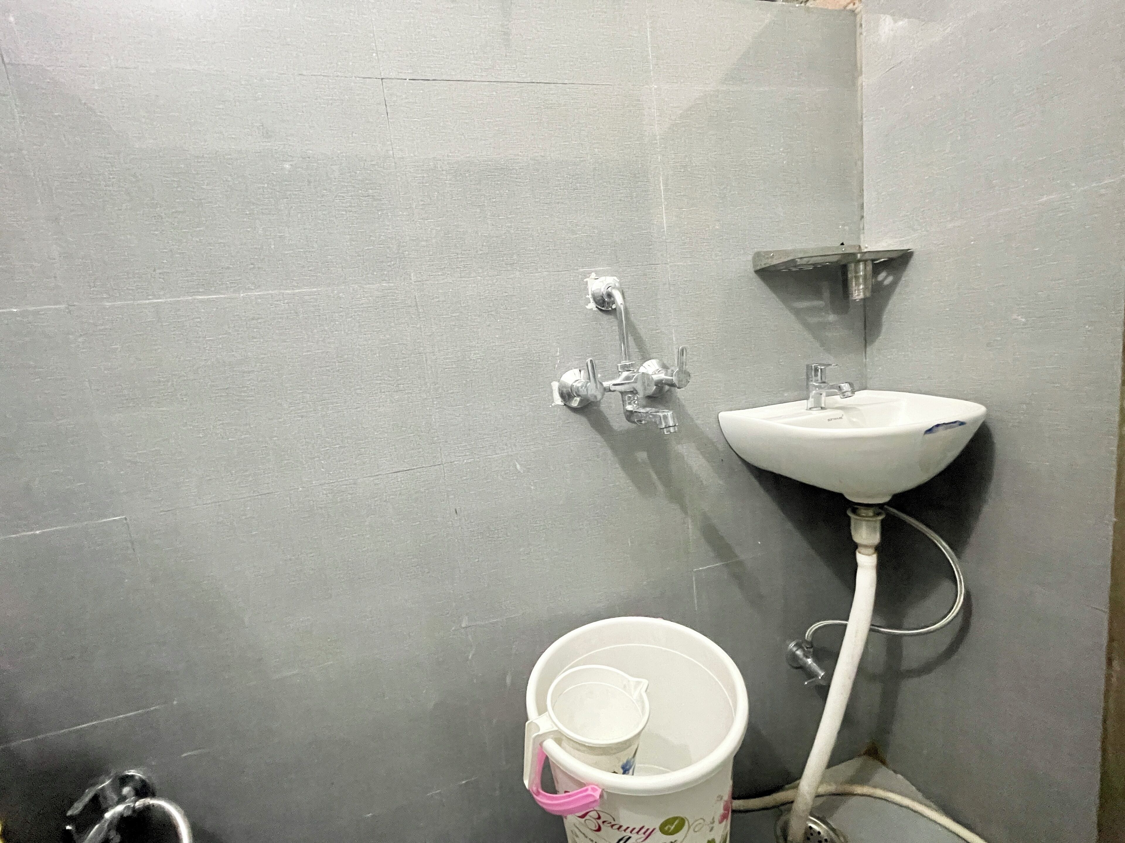 A bathroom with a grey tiled wall, a white sink, and a bucket with a pink handle.