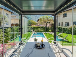 Ein Speisebereich mit Glaswänden und Blick auf einen Pool, mit Sitzgelegenheiten im Freien und einem Tisch, der mit Weingläsern und einer Flasche gedeckt ist.