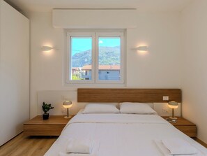 A bedroom with a wooden headboard, a large window with a view of houses and hills, bedside lamps, and a bedside table with a plant. - Allegra by Interhome (Dervio)