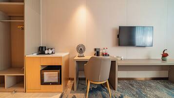 Habitación de hotel pequeña y funcional con escritorio, silla, televisor y una kitchenette.