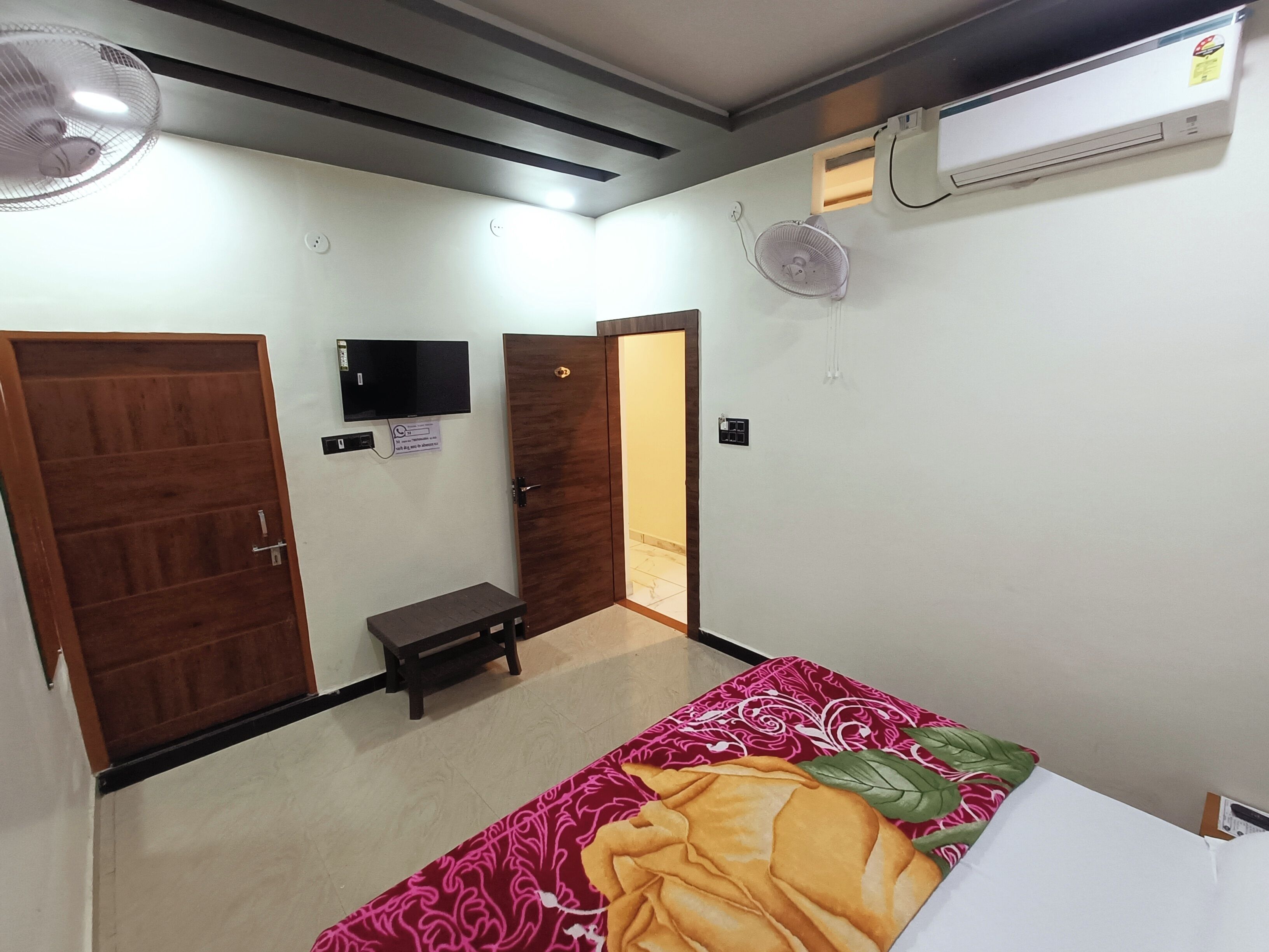 A hotel room with a bed, two doors, a small table, a TV, an air conditioner, and a ceiling fan.